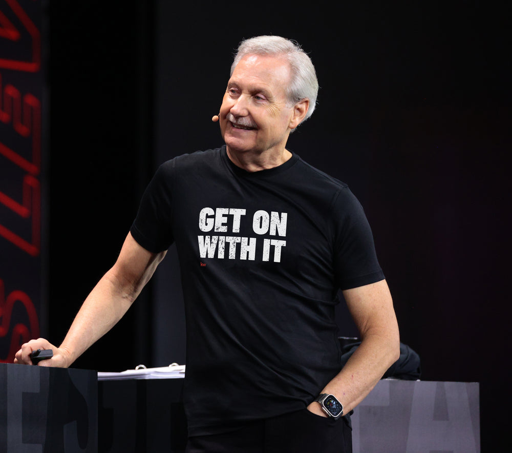 Gary Keller wearing a black t-shirt with 'GET ON WITH IT' text, standing against a dark background.