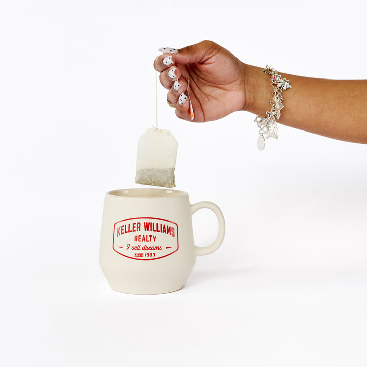 Hand placing a tea bag into a mug with 'Keller Williams Realty' branding on a white background